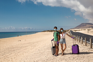 Young happy couple with surgery protective masks at  empty beach with suitcases during coronavirus summer holidays in Fuerteventura