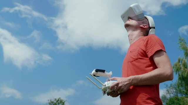 Man controls a quadrocopter through a remote control and looks at the video with goggles on his head. Drone operator in red T-shirt in a park shoots video on drone. Blue sky on the background