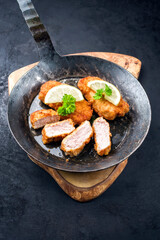 Traditional deep fried veal steak with lemon slices and herbs offered as closeup in a rustic old wrought iron skillet on a wooden board