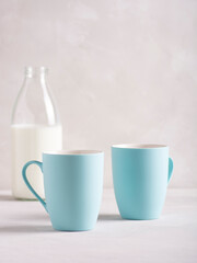 Milk in a glass bottle stands on a gray concrete table, next to it are two blue mugs. Vertical orientation, there is copy space.