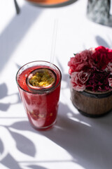 Red cold lemonade, decorated with a half passion fruit. A glass glass stands on a white tablecloth. Nearby is a wooden vase with red and pink cloves. A summer terrace. Alcoholic and non-alcoholic 