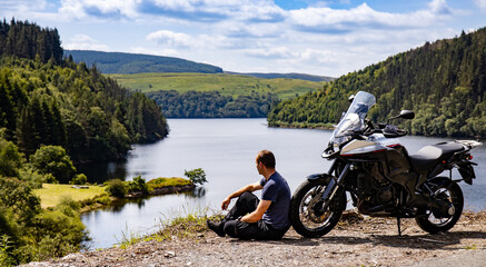 Naklejka premium Adventure motorcycle and biker man traveling, sitting and watching landscape with lake and mountains, freedom travel lifestyle in Wales UK