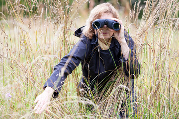 Blond hair mature woman spying with binoculars, move to the side dry grass, black clothing