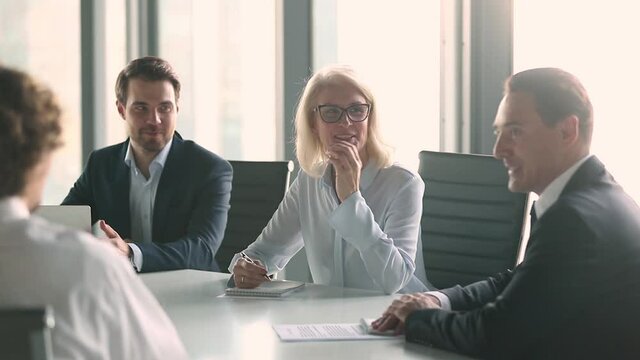 Successful well-dressed company male members headed by middle aged team leader woman in eyeglasses gathered together in boardroom taking part in briefing current matters negotiating sitting at desk..