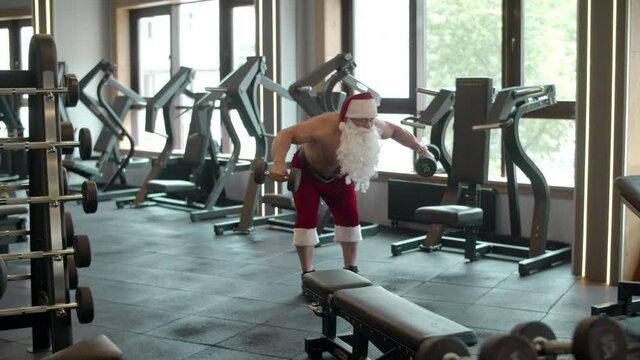 Santa claus doing dumbbell swings at gym. Man in santa costume in sport club