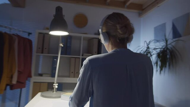 360-degree tracking shot of beautiful blonde woman in wireless headphones listening to music on smartphone with pleasure and moving her head to rhythm while working in design studio at night