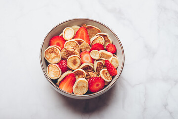 Trendy home breakfast with tiny cereal pancakes with piece of butter and fresh strawberries in the bowl. View from above.