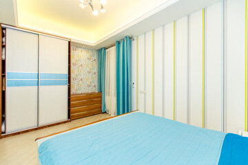 A bright bedroom with a large white double bed with a turquoise bedspread. turquoise drapes on the window. Light Wallpaper with vertical stripes. 