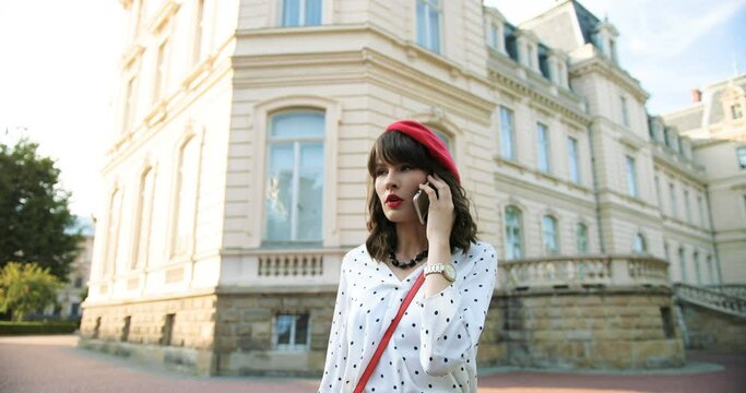 Portrait of pretty stylish girl in red beret chatting on cellphone while walking on street. Beautiful Caucasian young female with red lips talking on smartphone and walking outdoors. Call concept