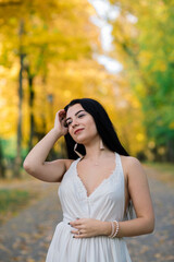 Young beautiful brunette female in a long white dress in autumn park.