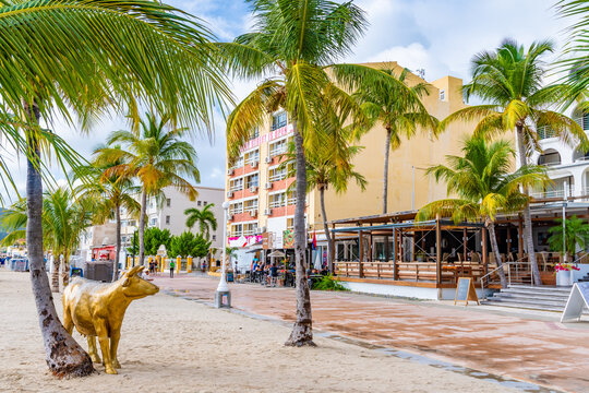Philipsburg, Sint Maarten. Holland House Beach Hotel golden cow on sand below coconut palm trees by waterfront coast with paved boardwalk and buildings in the Caribbean island city.