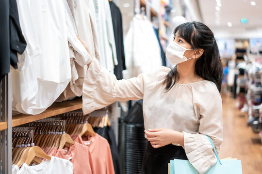 Asian woman wearing mask over her face while choosing shirt at shopping mall with shopping bag for healthcare and prevention from coronavirus, Covid19 influenza in crowded place..