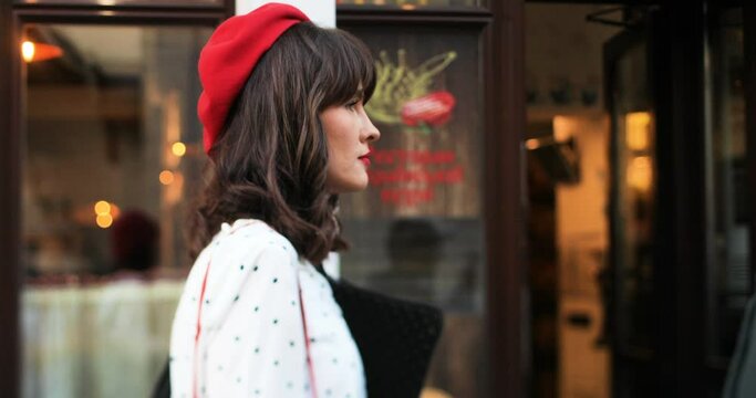 Side view of Caucasian pretty young stylish woman walking on street in crowded town. Close up portrait of beautiful girl in red french beret walking outdoors. Fashion lifestyle. Urban concept