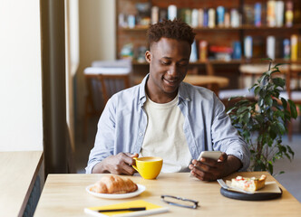 Fototapeta na wymiar Attractive African American guy using smartphone white having breakfast in cafe