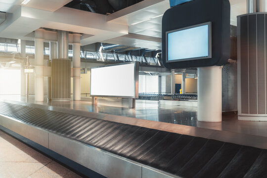 A baggage claim area in a hall of a modern airport arrival zone with conveyor belt and a mock-up of white empty advertising or information billboard on a chrome surface, bluish tv plasma screen above