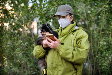 A girl wearing respirator mask and dialing a smartphone walking with a dog in the forest at COVID-19 period.