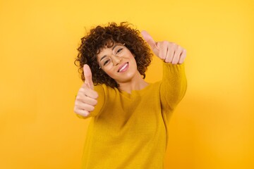 Young beautiful business woman with curly hair wearing yellow sweater approving doing positive gesture with hand, thumbs up smiling and happy for success. Winner gesture.