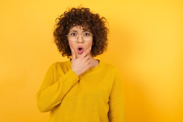 Young beautiful business woman with curly hair  wearing yellow sweater standing over white isolated background Looking fascinated with disbelief, surprise and amazed expression with hands on chin