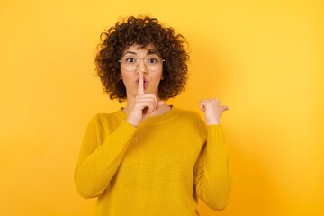 Young beautiful African American woman with curly hair wearing yellow sweater asking to be quiet...