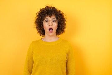 Oh my God. Surprised  arab female stares at camera with shocked expression, wears yellow sweater, exclaims with unexpectedness, isolated over yellow background