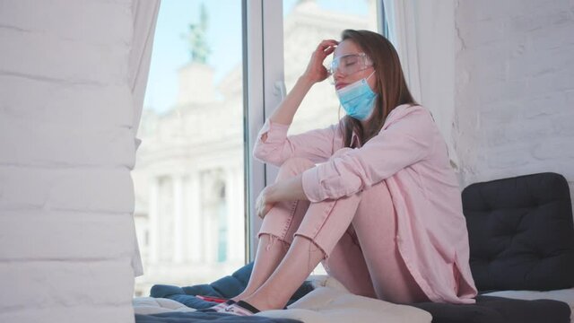 Pensive upset caucasian woman taking off medical mask wearing protective eyeglasses sitting in despair by window. Depression. Coronavirus. Lockdown stress. Self-isolation.