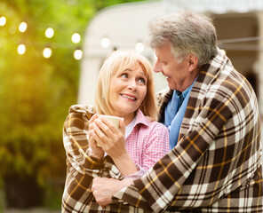 Camping romance. Affectionate mature couple cuddling under warm plaid near trailer outside