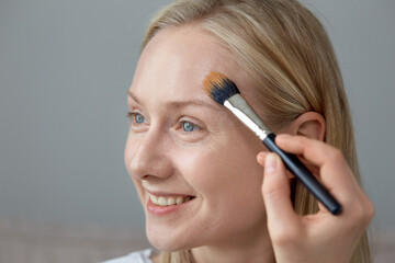 Fototapeta premium A smiling young girl puts a tonal cream on her face with a brush.