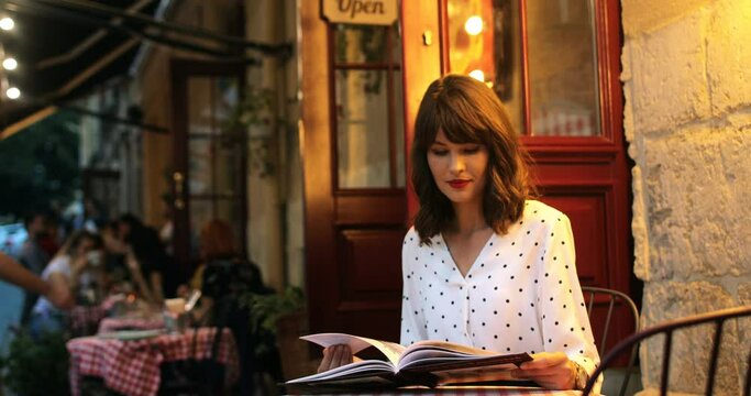 Portrait of beautiful charming paris girl reading magazine while sitting in cafeteria outdoor. Joyful pretty Caucasian woman resting in cozy restaurant and reading book. Leisure concept