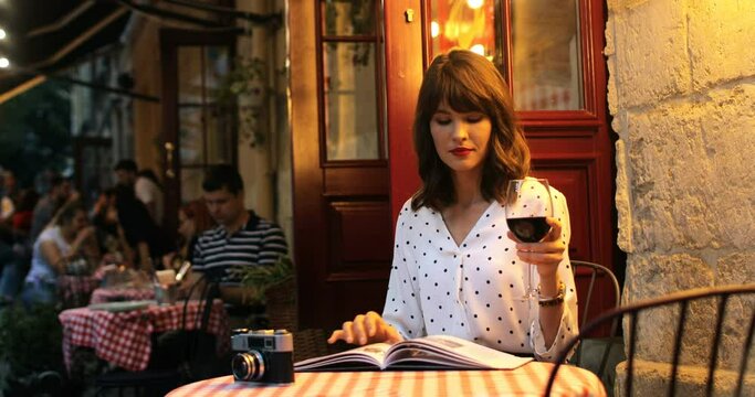 Portrait of beautiful charming girl reading fashion magazine and drinking wine while sitting outdoors. Joyful pretty Caucasian woman resting in cozy restaurant with glass of red wine and reading book.