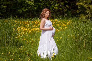 Fototapeta premium Beautiful pregnant woman in white dress standing in the green park in sunny day.