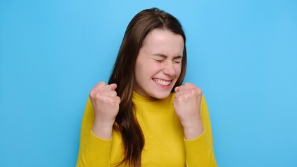 Triumphing ambitious young woman celebrates amazing victory with both clenched fists, rejoices over awesome achievement, exclaims positively hooray, wears sweater, isolated on blue studio background