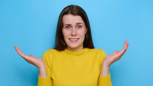 Portrait of brunette woman with puzzled expression, spreads hands with confused look, frowns face, feels hesitant, indignant, wears yellow sweater, isolated over blue studio background. Doubt concept