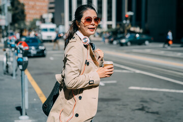 Side portrait of smiling asian korean woman in spring coat walking in modern city with headphones and coffee cup. beautiful office lady standing on road looking aside to wait for time to cross road