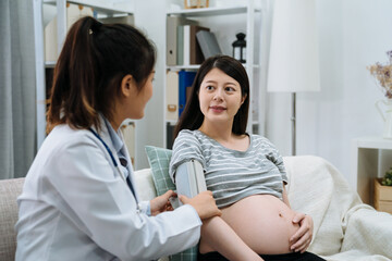 Fototapeta premium female asian doctor measuring blood pressure of pregnant woman in home visit. elegant maternity lady with naked abdomen sitting on couch in apartment living room. midwife doing health check of mom