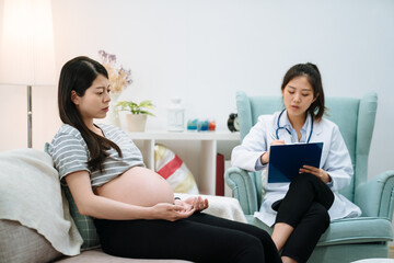 asian chinese pregnant woman visiting psychologist doctor in clinic. female psychological consultant talking and giving advice writing on clipboard. frowning upset future motherhood sitting on couch