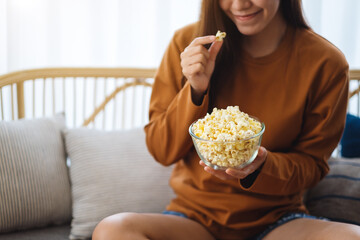 Close up image of a beautiful young woman holding and eating pop corn while sitting on sofa at home