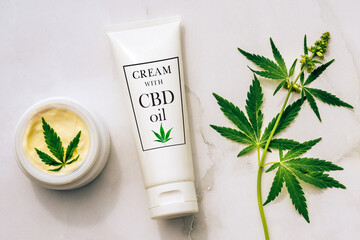 Tuba and jar of cream CBD oil, THC tincture and hemp leaves on marble background. Flat lay,...