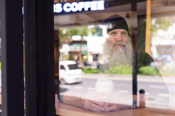 Mature handsome bearded hipster man thinking at the coffee shop through the window