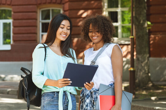Two multicultural female students standing outdoors near university campus with workbooks
