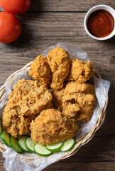 Crispy fried chicken in wicker basket serve with cucumber, tomato and ketchup on wooden background. top view. Flat lay.