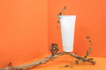 One white blank cosmetic tube bottle on wooden stick with dried flowers and eucalyptus branch in corner space on orange background. Mockup Front view Copy space