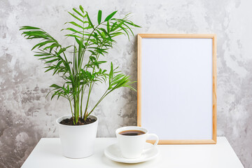 Wooden vertical frame with white blank card, cup of coffee and green exotic palm flower in pot on table on gray concrete wall background. Mockup Template for your design, text