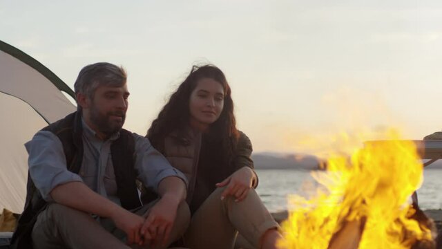 Cinemagraph of romantic tourist couple sitting together beside tent on lakeshore and looking at campfire