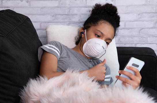 Female Child Wearing Protective Mask,  Using Mobile Phone At Sofa, Show Thumb Up Sign During Video Conference With Mobile Phone. Corona Virus Concept.
Video Call.