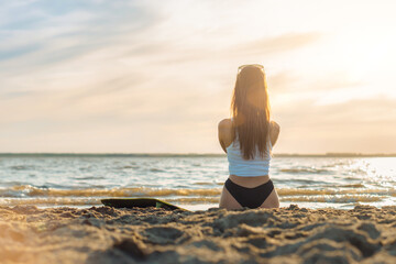 Fototapeta na wymiar Young caucasian woman surfer in white shirt sitting on the beach with her surfboard watching the sun rise or sun set. Summer concept
