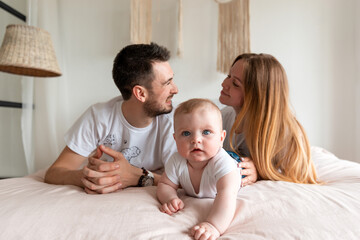 Fototapeta na wymiar Happy family: mother, father and daughter on a white bed in a sunny bedroom. Parents and a small child are resting at home.