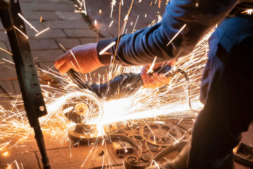 Side view of a man's hands working on a metal part of a garden bench, using an electric grinder while sparks are flying around in the industrial workshop.