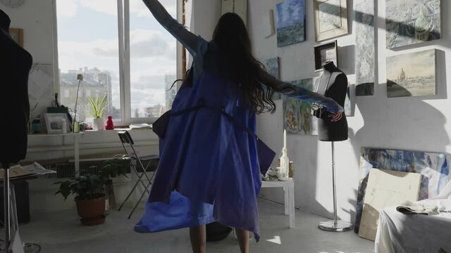 positive barefoot ballerina in stylish loose blue clothes dances in modern art studio with pictures at sunlight slow motion