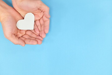 Cupped hands holding a white heart in blue background. Charity, pure love, compassion and kindness concept. Top view.