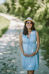 Outdoor portrait of happy pregnant woman with sunglasses and hat - summer time in nature - beautiful moments of family life